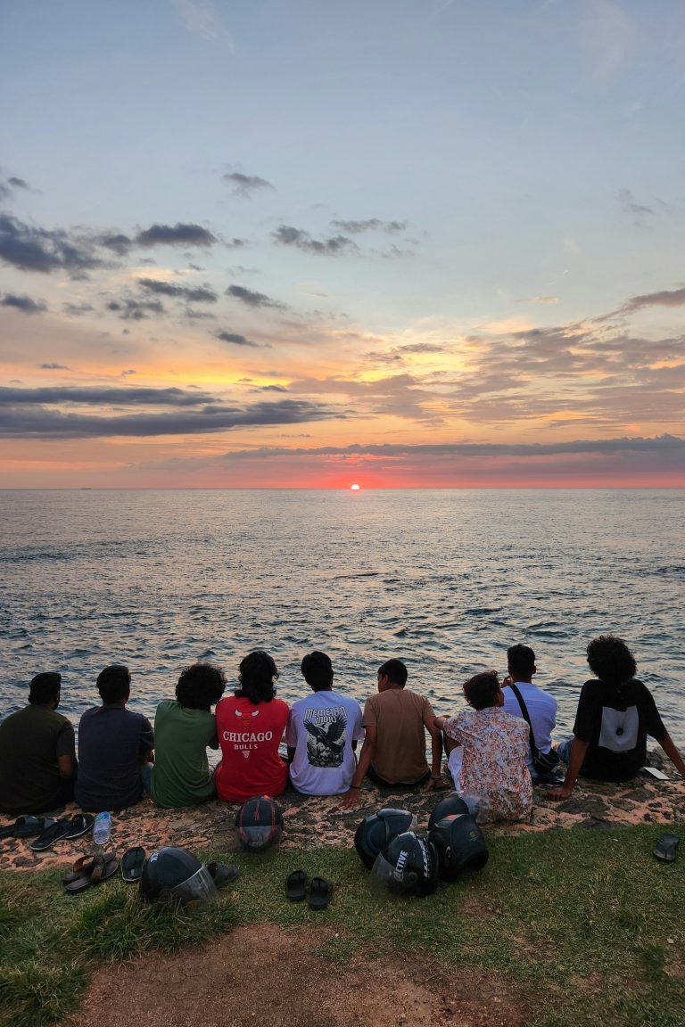 Een groep mensen op een kleinschalige groepsreis zit op een randje met uitzicht op de oceaan en kijkt naar de zon die ondergaat aan de horizon met kleurrijke wolken in de lucht.